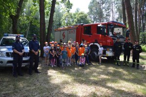 Dzieci z policjantami i strażakami na polanie w lesie obok stojącego radiowozu i wozu strażackiego