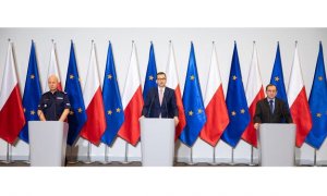 Na zdjęciu od lewej: gen. insp. Jarosław Szymczyk- Komendant Główny Policji, Premier Mateusz Morawiecki i minister MSWiA Mariusz Kamiński. W tle flagi Polski i Unii Europejskiej.