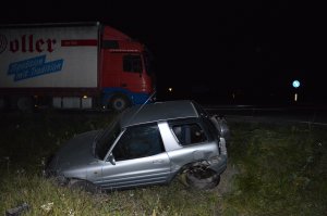 Uszkodzony samochód koloru szarego znajdujący się w rowie przy drodze, w tle samochód ciężarowy.