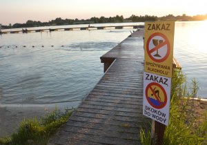 Drewniany pomost nad wodą. Przed nim znak zakazu spożywania alkoholu i znak zakazu skoków do wody