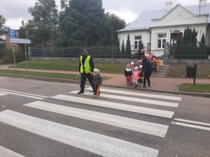 Policjant z dzieckiem na przejściu dla pieszych, za nimi dzieci z opiekunkami. W tle budynek.