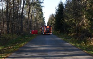 Wóz bojowy straży pożarnej na drodze biegnącej przez las. W tle czerwony parawan, a za nim rozbity samochód.