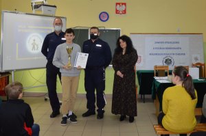Policjanci, nauczycielka i uczeń, który trzyma w rękach puchar i dyplom w sali lekcyjnej. W tle dwie tablice informujące o zawodach i konkursie.