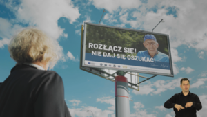 Zdjęcie przedstawia starszą kobietę patrzącą w bilbord na którym jest inny starszy mężczyzna i napis Rozłącz się nie daj się oszukać.