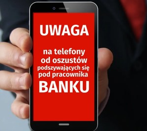 telefon komórkowy w dłoni i biały napis na czerwonym tle na ekranie 
&quot;Uwaga na telefony od oszustów podszywających się pod pracownika banku&quot;.