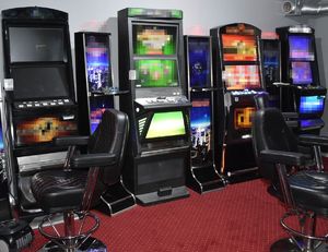 Cztery stojące w pomieszczeniu automaty do gier hazardowych, obok nich fotele do siedzenia.