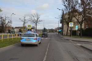 Radiowóz na ulicy przed przejściem dla pieszych, a za przejściem dla pieszych samochód osobowy.