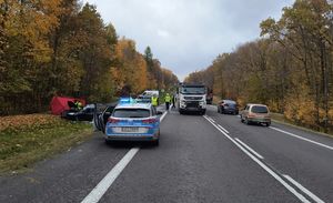 Radiowóz na drodze oraz samochód ciężarowy, a w tle policjanci wykonujący czynności na miejscu śmiertelnego wypadku.