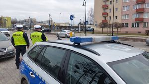 Policjanci stoją przy radiowozie i obserwują zachowania uczestników ruchu drogowego w rejonie przejścia dla pieszych.