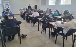 Uczniowie przy stolikach rozwiązują test wiedzy, a przy biurku stoi policjantka.