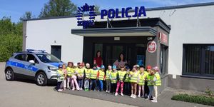 Grupa przedszkolaków wraz z wychowawczyniami pozuje przed budynkiem posterunku, a obok stoi zaparkowany radiowóz.