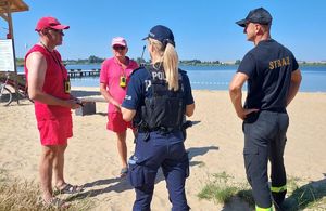 Policjantka ze strażakiem rozmawiają z dwoma ratownikami wodnymi nad zalewem.