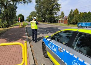 Policjant prowadzi pomiar prędkości pojazdu na drodze. Za nim stoi radiowóz.