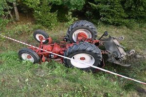 Leżący w rowie na trawie przewrócony do góry kołami ciągnik rolniczy.