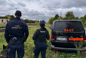 Policjantka i funkcjonariusz Straży Ochrony Kolei wraz z psem służbowym obserwują przejazd przez torowisko. Obok stoi zaparkowany pojazd służbowy Straży Ochrony Kolei.
