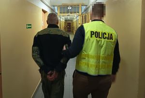 Policjant w odblaskowej kamizelce z napisem Policja prowadzi korytarzem w budynku komendy zatrzymanego mężczyznę.