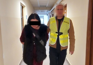 Nieumundurowany policjant w odblaskowej kamizelce z napisem Policja prowadzi zatrzymaną kobietę