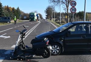 Uszkodzony samochód i motorower na jezdni. W tle policjant kierujący ruchem na drodze.