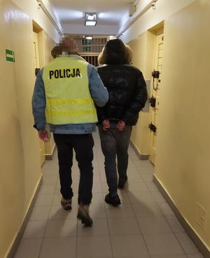 Zdjęcie przedstawiające funkcjonariusza Policji, który prowadzi osobę zatrzymaną. Policjant ma na sobie żółtą kamizelkę.