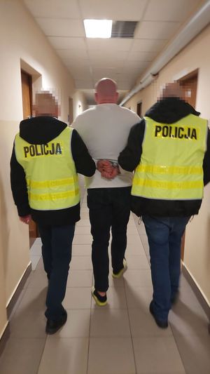 Zdjęcie przedstawiające dwóch policjantów w żółtych kamizelkach prowadzących osobę zatrzymaną