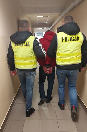 zdjęcie przedstawiające dwóch funkcjonariuszy w żółtych kamizelkach prowadzących osobę zatrzymaną. Mężczyzna ubrany w bluzę koloru czerwonego oraz spodnie koloru niebieskiego